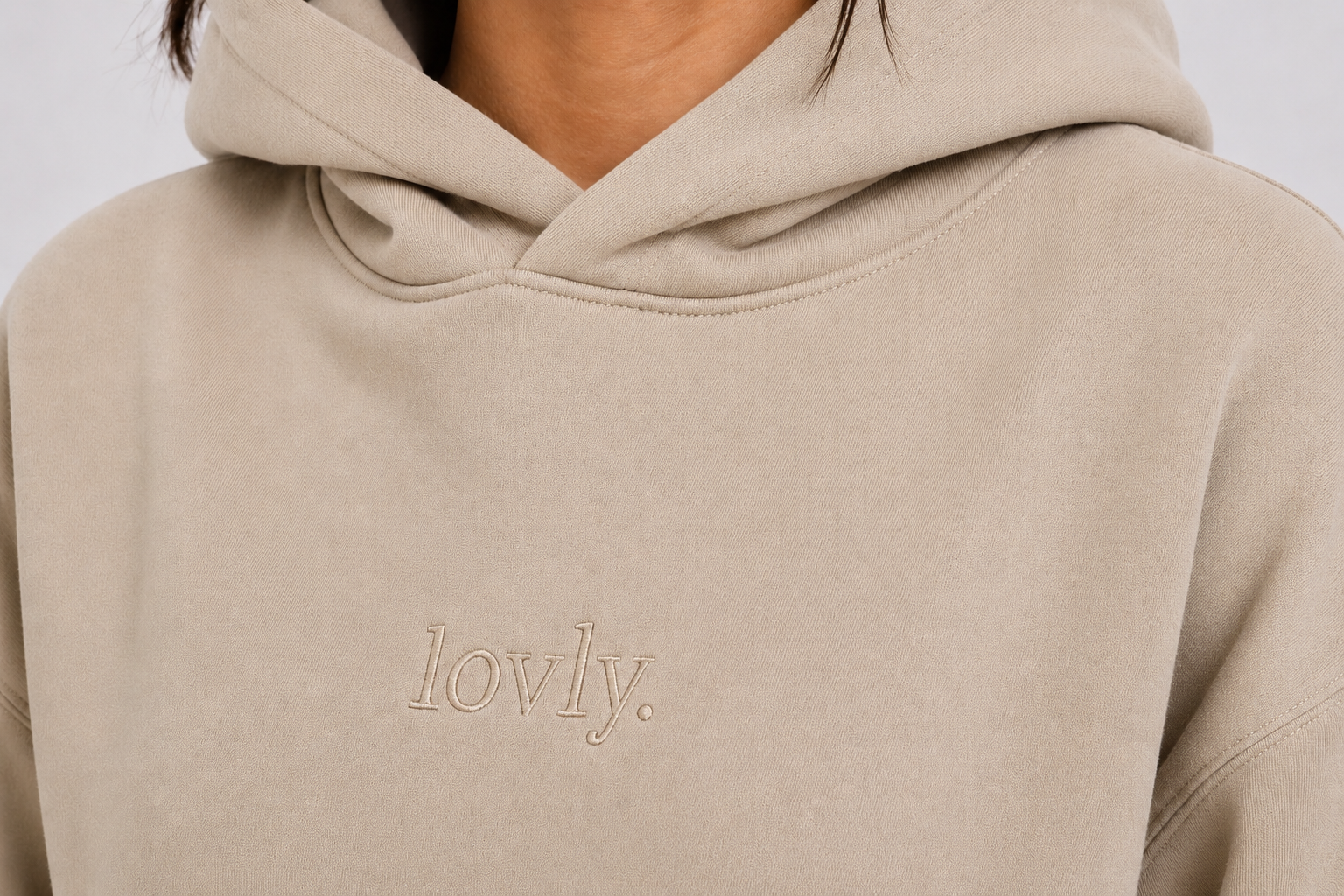 Close-up of a person wearing a light beige hoodie with the word 'lovly.' embroidered on the chest.